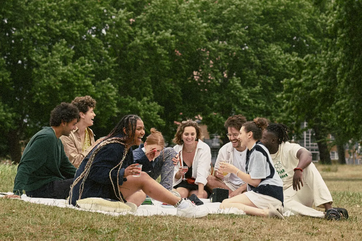 Friends laughing together on a picnic blanket in a park, with tall trees and a tower block behind them